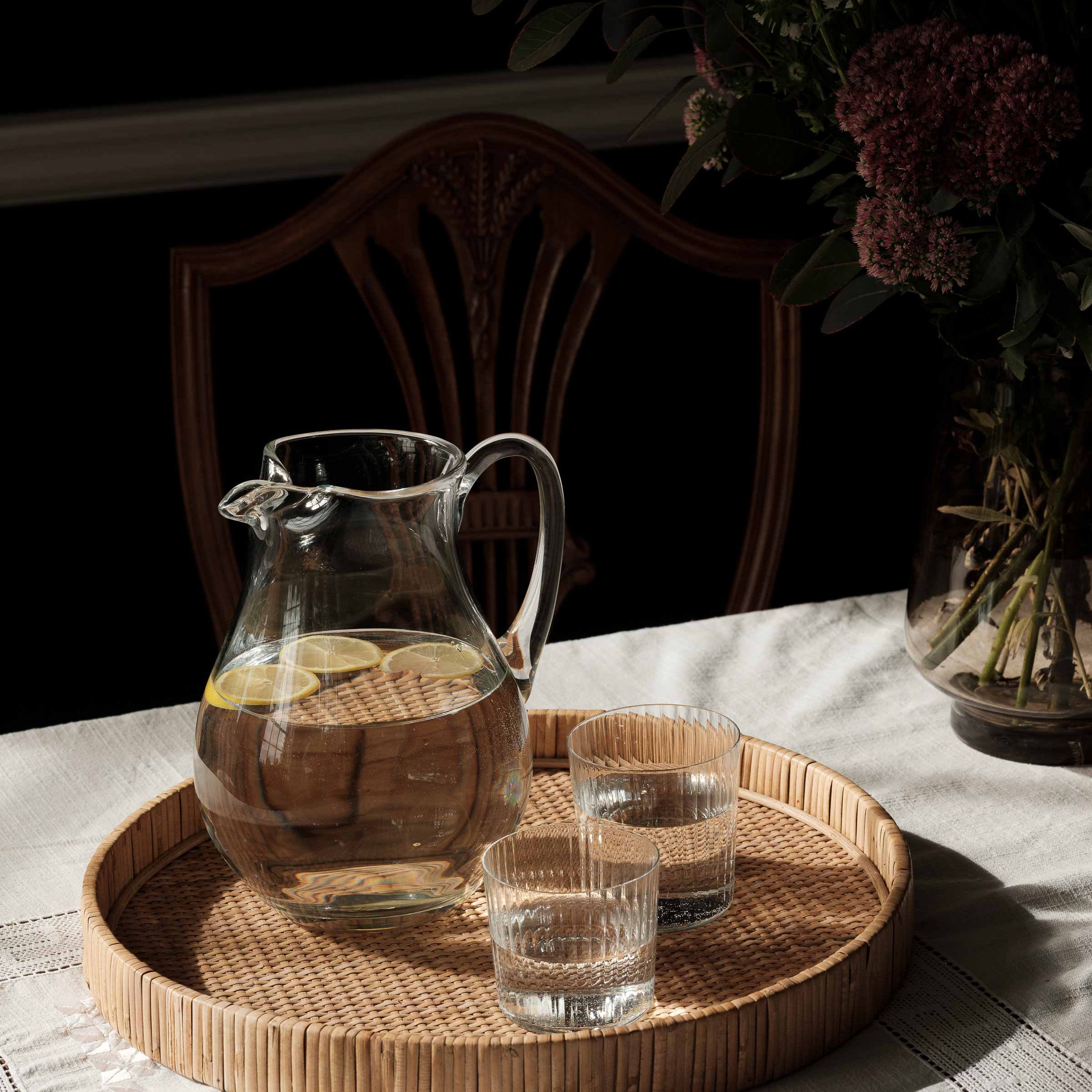 A set of four clear glass lowball glasses with a linear texture, filled with water and placed on a woven tray on a table, with a floral arrangement in the background.